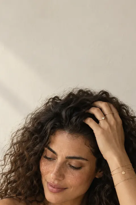 A woman with long curly hair smiling down, one hand in her hair, against a pale plaster wall
